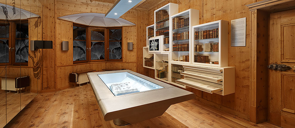 Bibliothek des Pharmaziemuseums Brixen. Wandschrank und "trickreicher Tisch" für wechselnde Ausstellungen, Entwurf Walter Angonese. Tischabdeckung aus rotem Kunstleder. Gesponsert von der Raiffeisenbank Brixen. Produziert von Barth Innenausbau. Holzgetäfel aus dem 17. Jh.